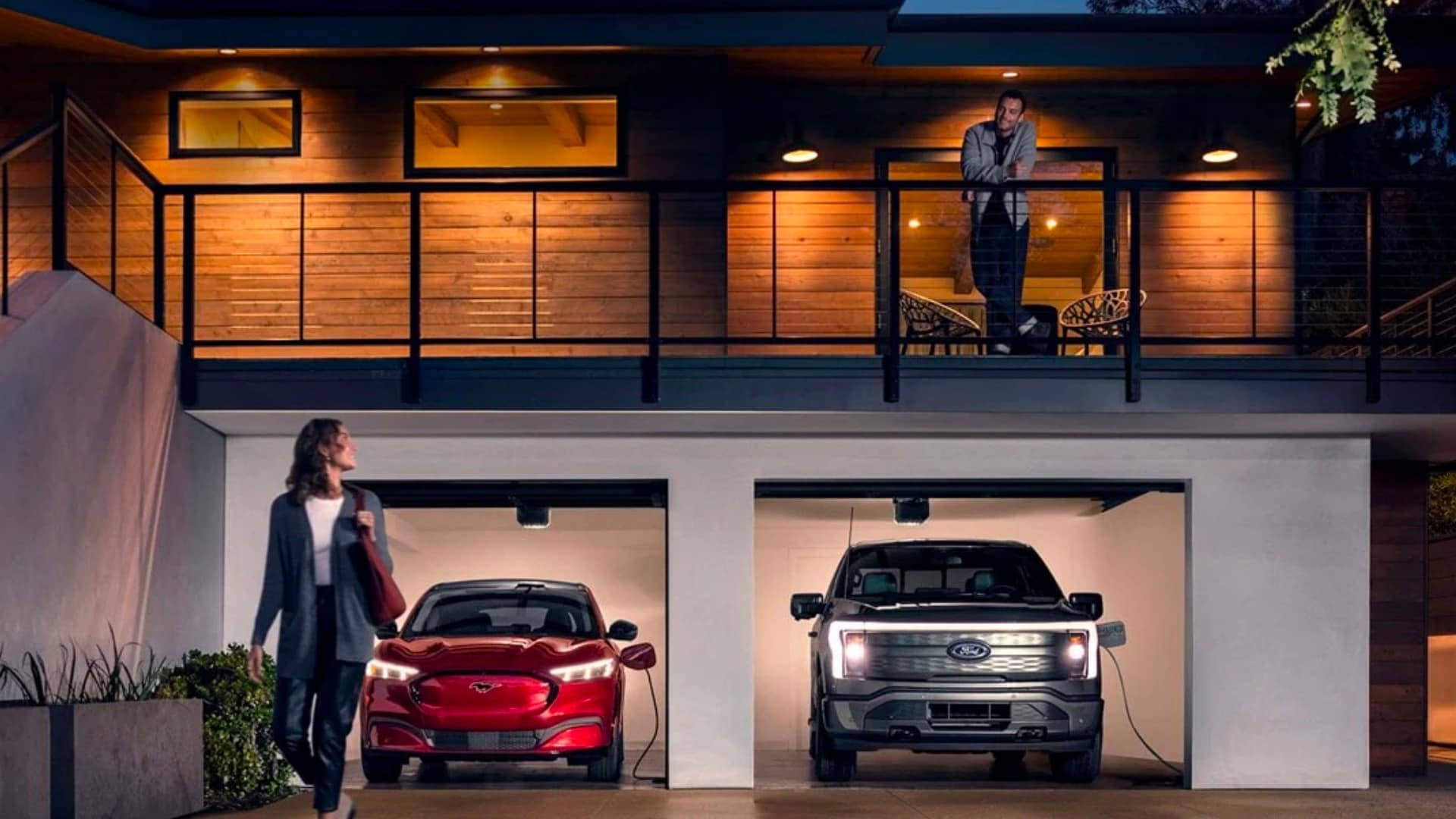 A 2022 Ford Mustang Mach-E and a Ford F-150 Lightning are being charged in a garage at night while a man stands on a balcony looking at awoman below, who is looking back at the man.