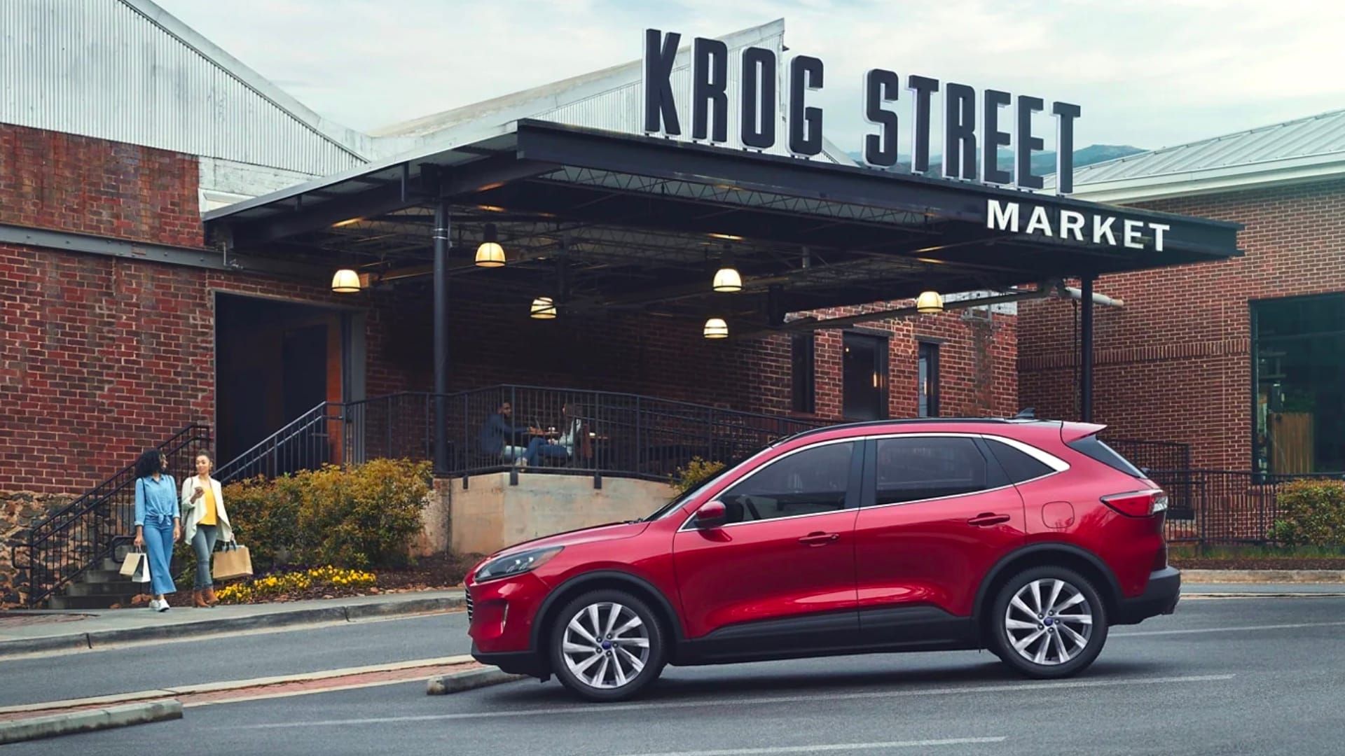 Learn about the differences between Hybrid and Electric vehicles Two women walk toward a Red Ford Escape parked outside of a building with a sign that says KROG STREET MARKET