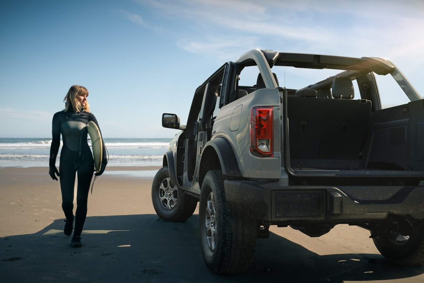 An image of a Ford Bronco parked on a beach next to a woman with a surf board.