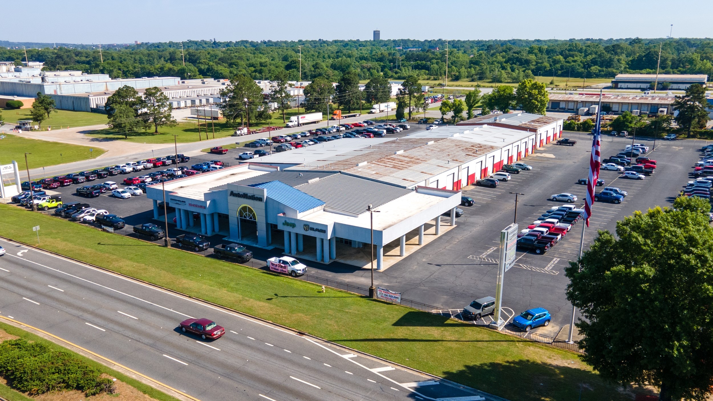 Overhead exterior view of AutoNation Chrysler Dodge Jeep Ram South Columbus during the day. There is a clear sky and many vehicles parked near the grey building. Trees and greenery are visible around the parking lot and surrounding area.