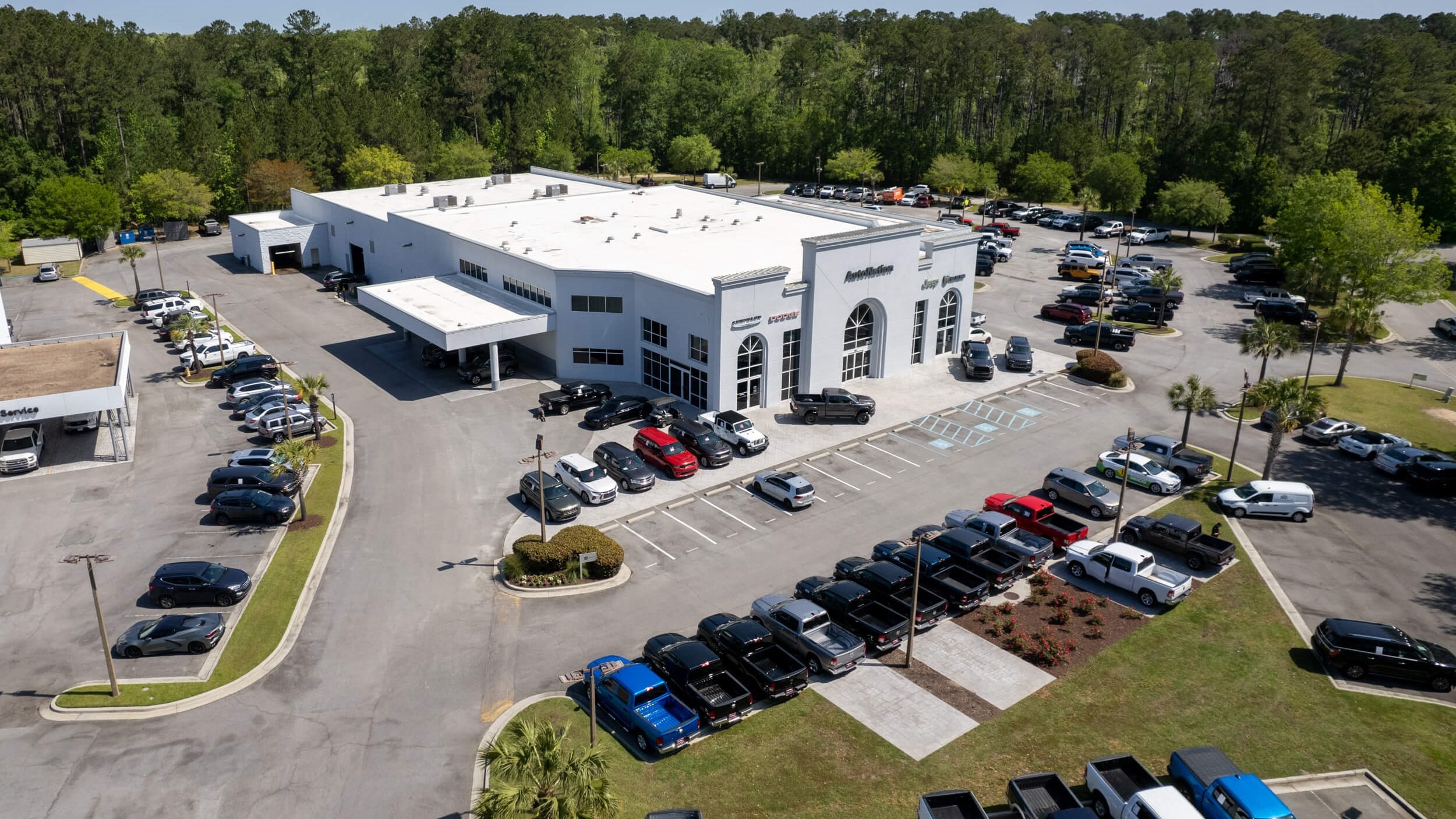 Overhead exterior view of AutoNation Chrysler Dodge Jeep Ram Hilton Head during the day. There is a clear sky and many vehicles parked near the grey building. Trees and greenery are visible around the parking lot and surrounding area.