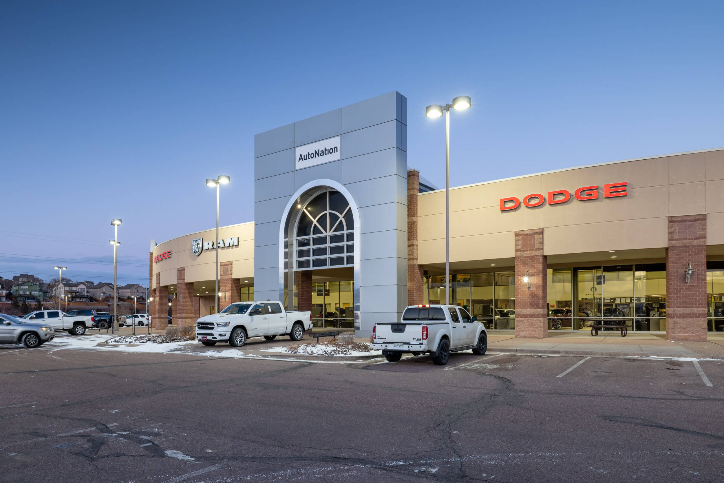 Exterior view of AutoNation Dodge Ram Colorado Springs with some cars parked in front and some snow in the parking lot. Clear sky with and a low light time of day