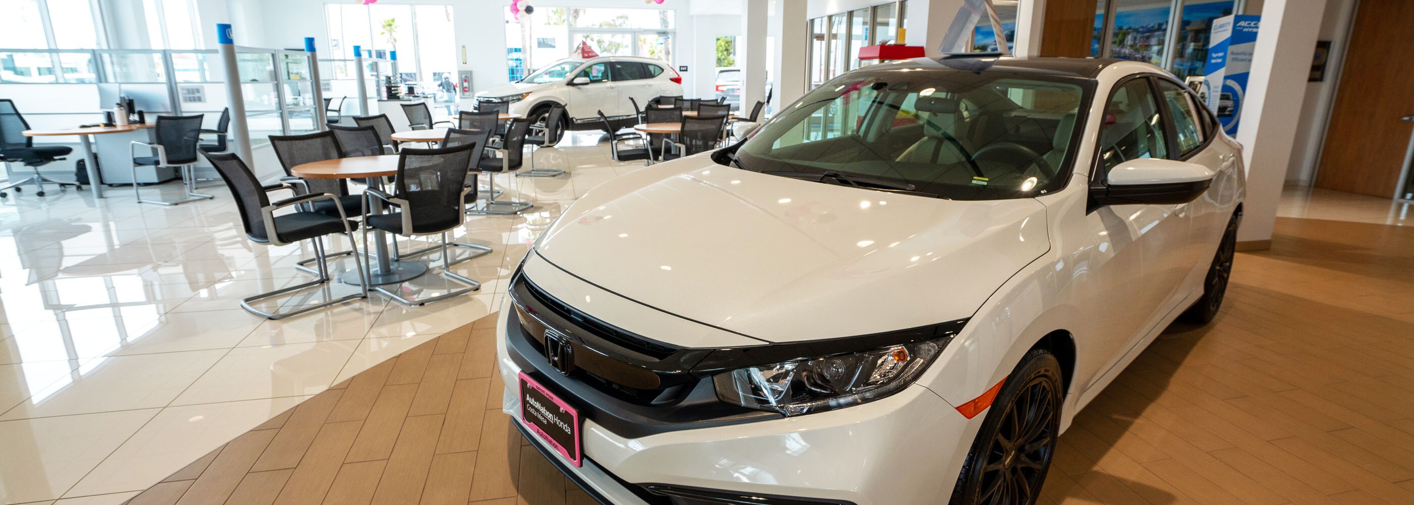 Interior view of AutoNation Honda Costa Mesa. A white Honda is parked with round tables and chairs nearby.