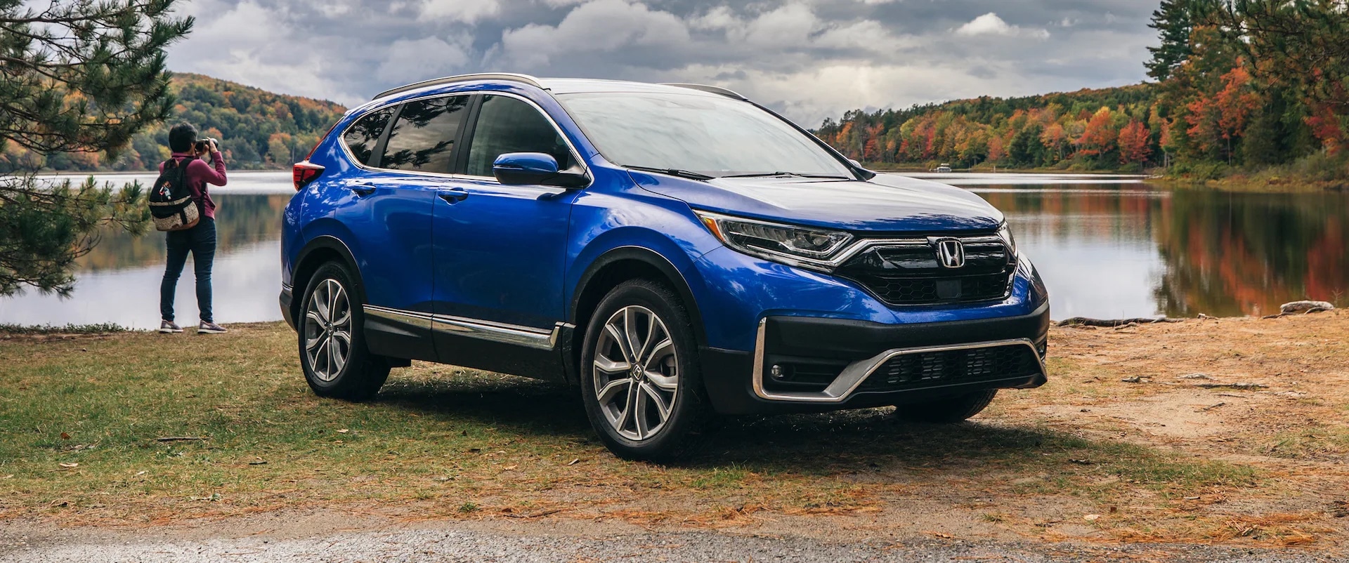 Blue Honda CR-V Hybrid parked alongside lake while a person stands near the vehicle and looks out at the lake.