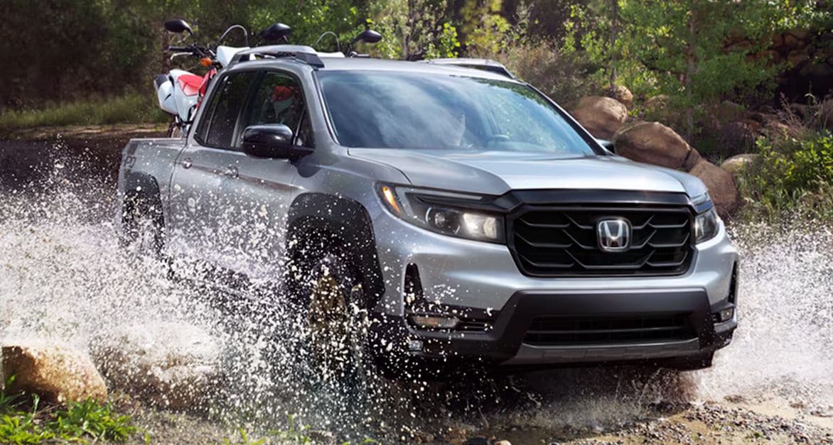 Honda Ridgeline in Lunar Silver Metallic Honda Ridgeline in Lunar Silver Metallic driving through a puddle of water in an off-road, forested trail during the day. Two dirtbikes are in the bed of the truck.