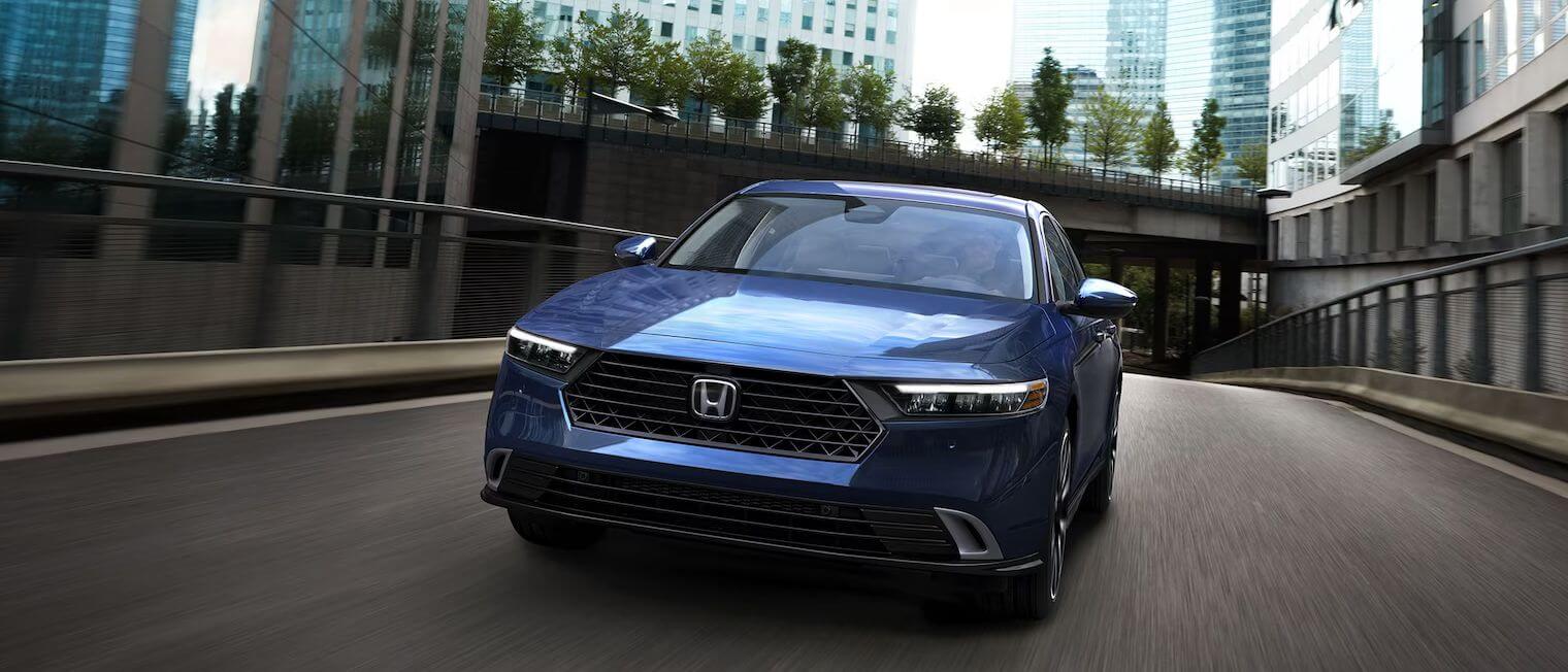 Honda Accord Hybrid in Canyon River Blue Metallic Exterior shot of a Honda Accord Hybrid in Canyon River Blue Metallic driving through a road that cuts through a downtown city setting.