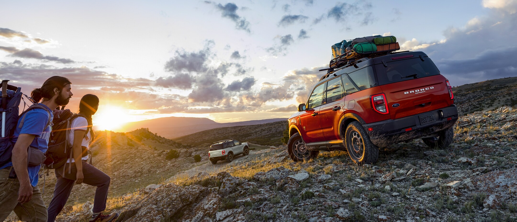 Two hikers walk towards a Red Ford Bronco Sport with luggage on the top. It sits on a rocky trail. Another white Ford Bronco is off in the distance.