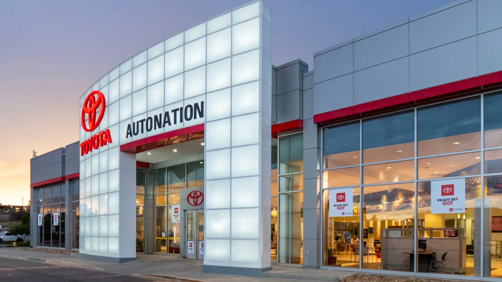 Exterior view of AutoNation Toyota Arapahoe, with either a sunset or a sunrise, as the sky in the background has some yellow and red. The building has a glass facade, and there are red and white posters on some of them.