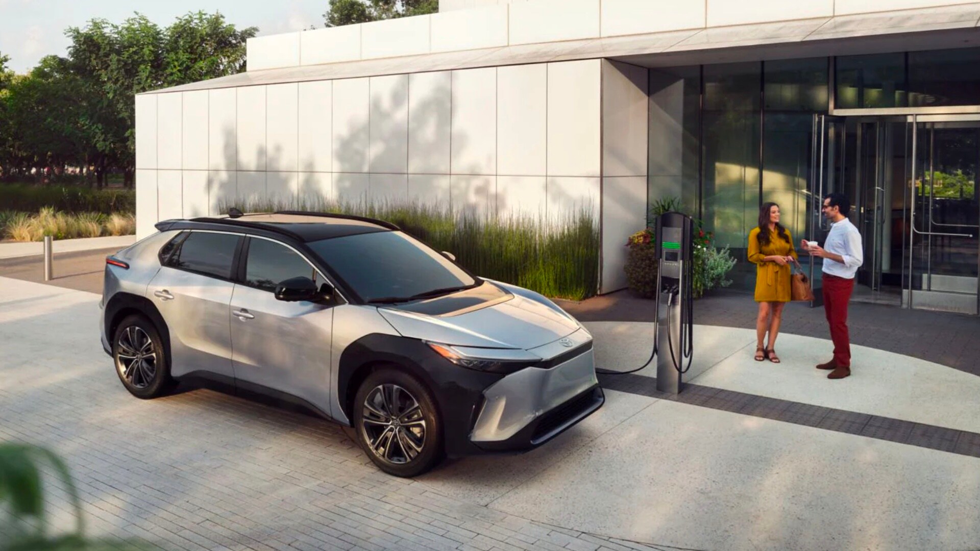 Electric Vehicles Buyers Guide Silver Toyota using charging station in front of modern looking one story building while a man and woman talk with each other in front of the building's entrance