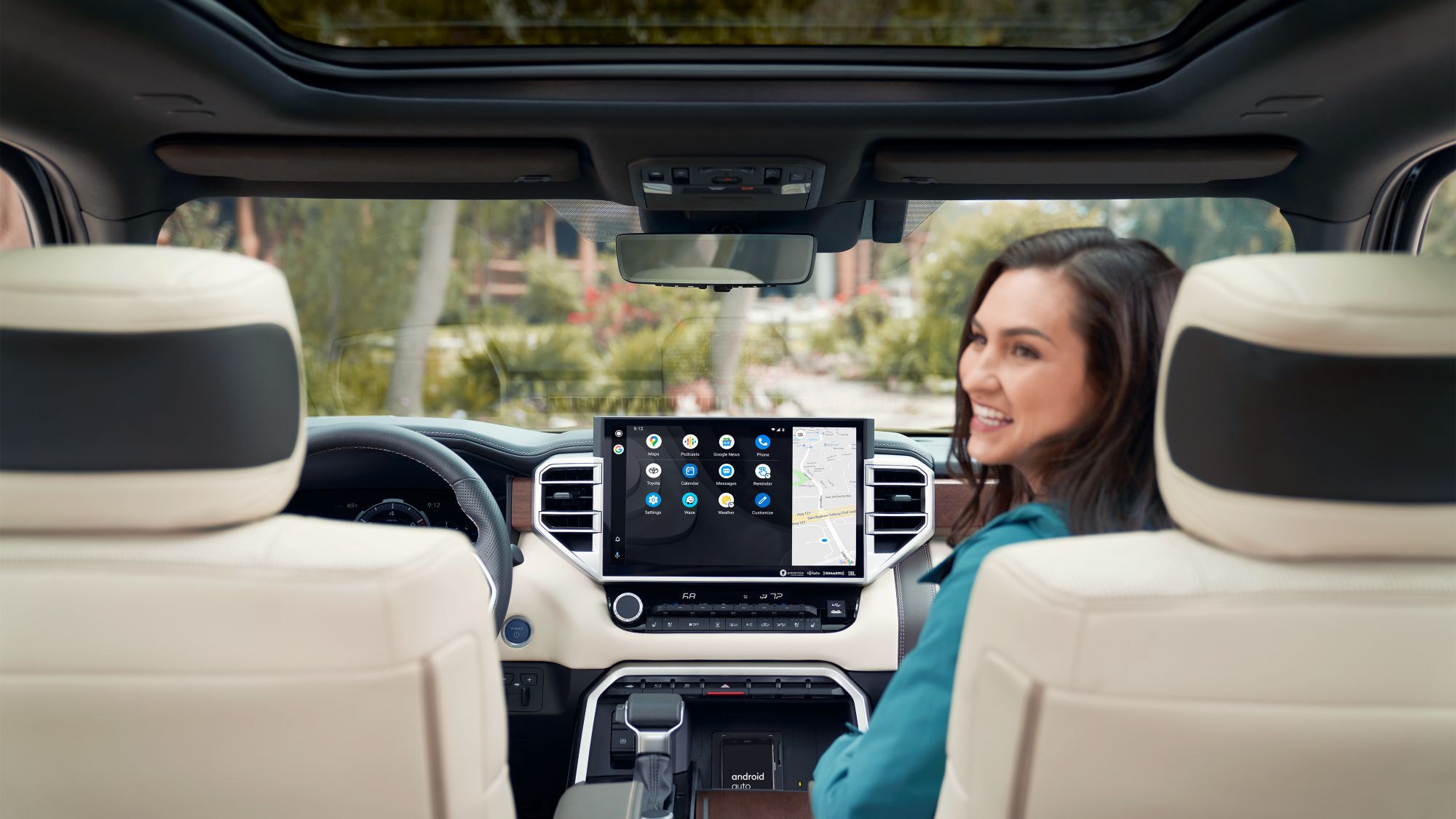 Interior view looking out the front of a Toyota from the backseat. A woman is sitting in the passenger seat and looking back. A touchscreen featuring the Android Auto UI is in the center of the frame.