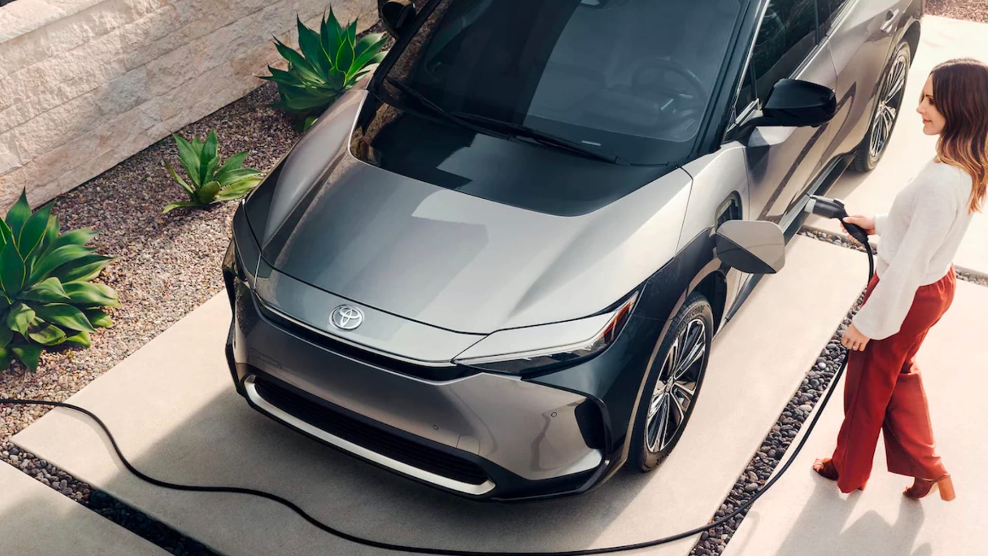 Charging Your Electric Vehicle Overhead view of a Silver Toyota with a woman wearing a white shirt and
red pants standing next to it and plugging in the charging cable