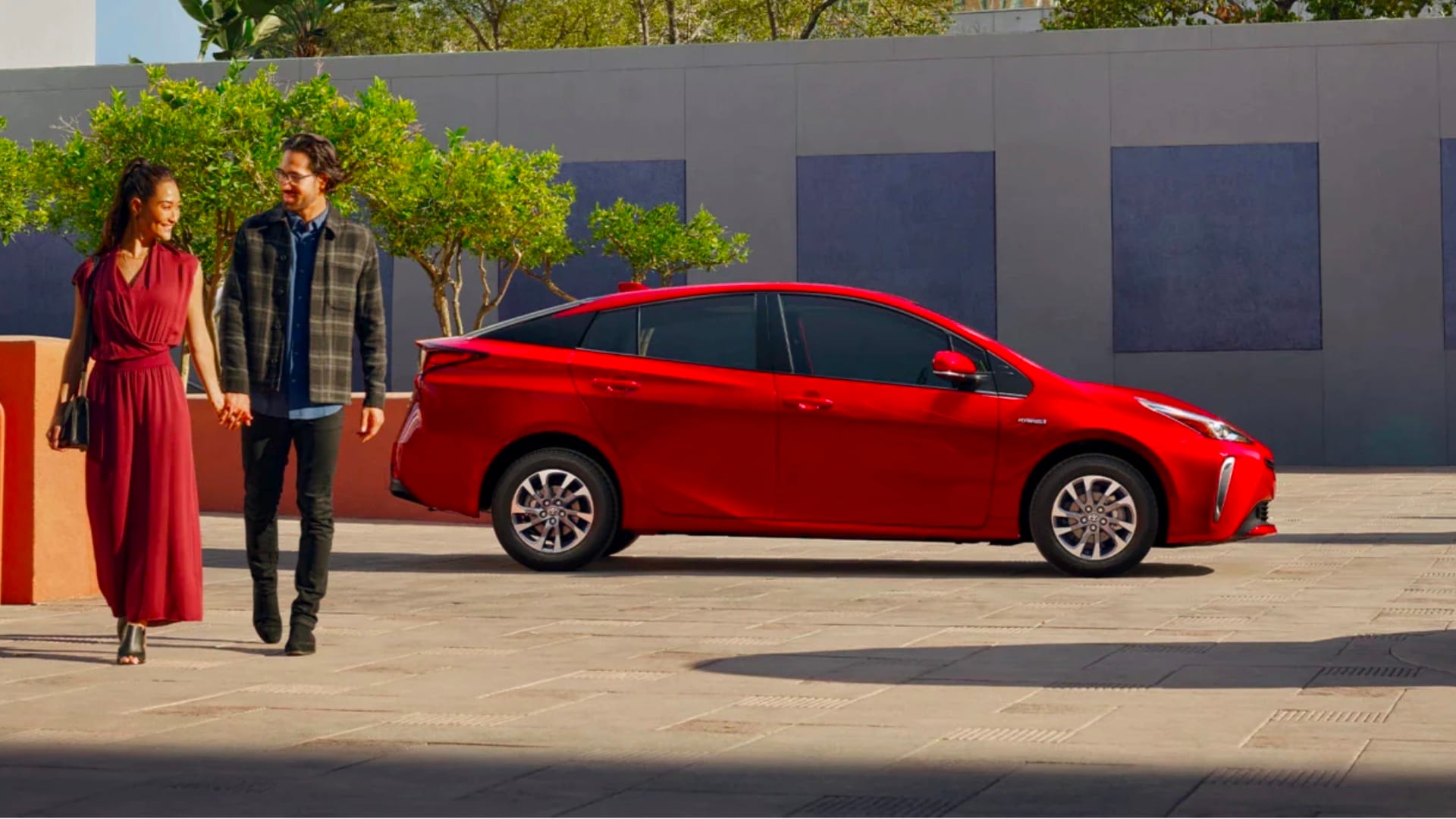 Hybrid vs Electric Vehicles Profile view of Red Toyota in background while two people walk next to each other towards camera