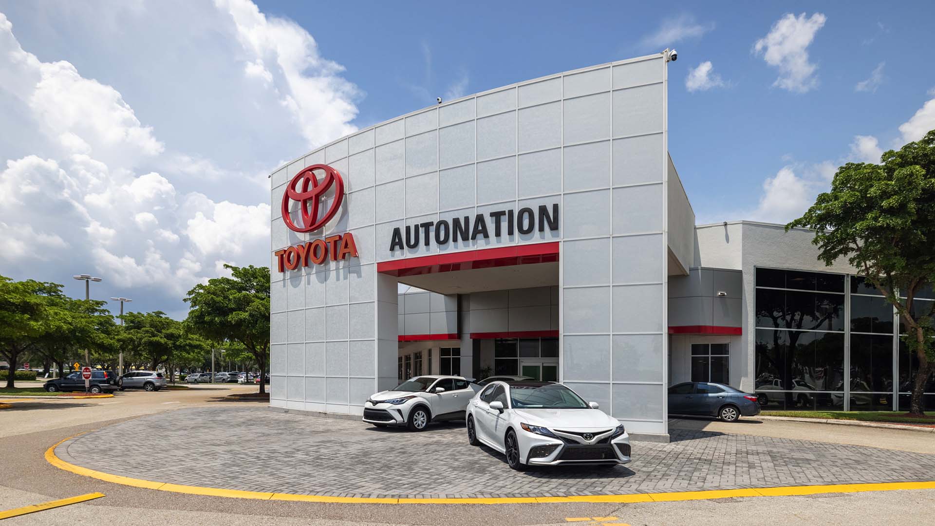 Exterior view of AutoNation Toyota Fort Myers, with a partly cloudy blue sky. The building is tall and grey and has some large windows. Several vehicles can be seen parked near the vehicle.