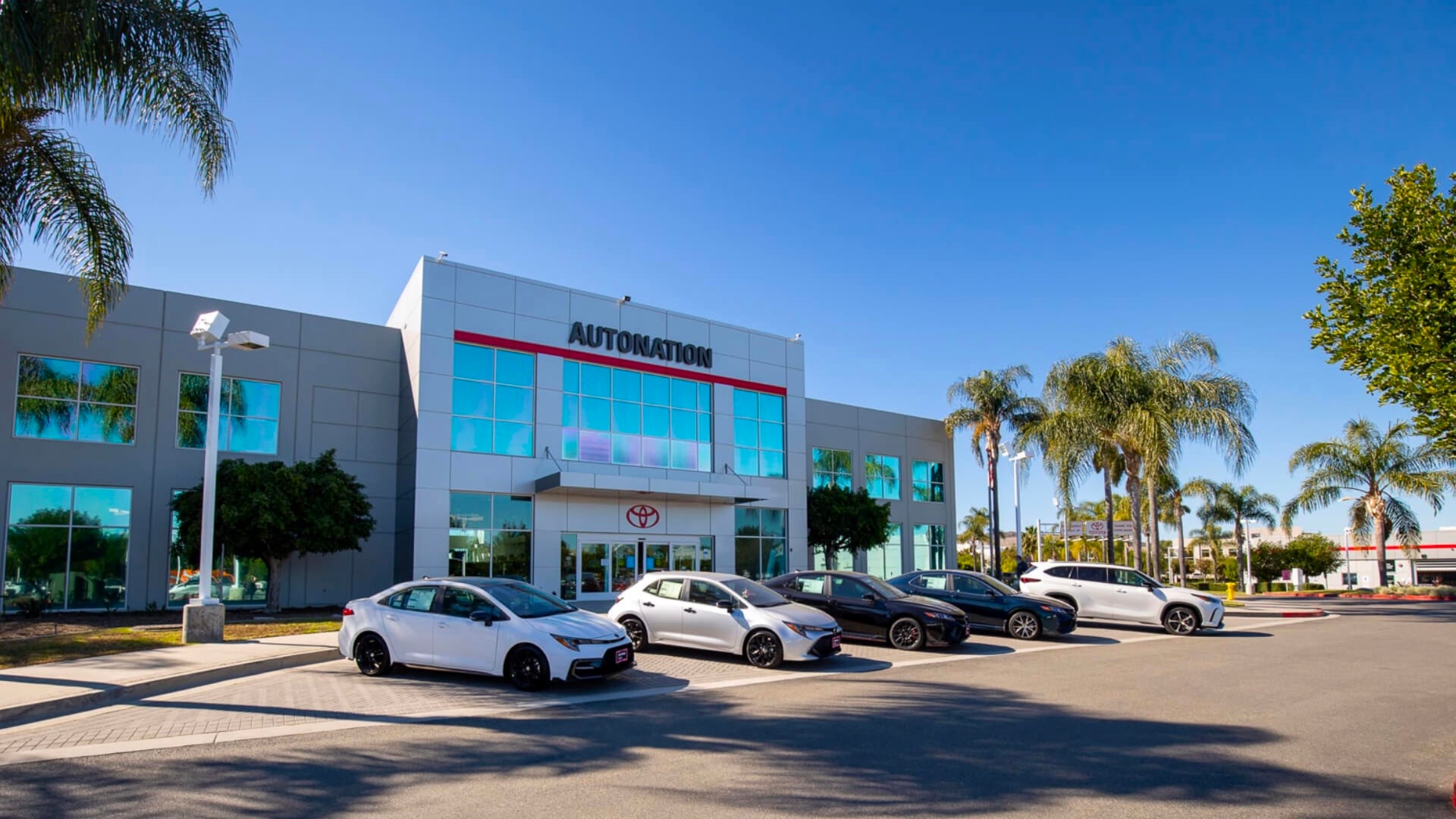 Exterior view of AutoNation Toyota Irvine. The building is grey and white and has some large windows. Many vehicles can be seen parked near the building, which has several palm trees anearby.