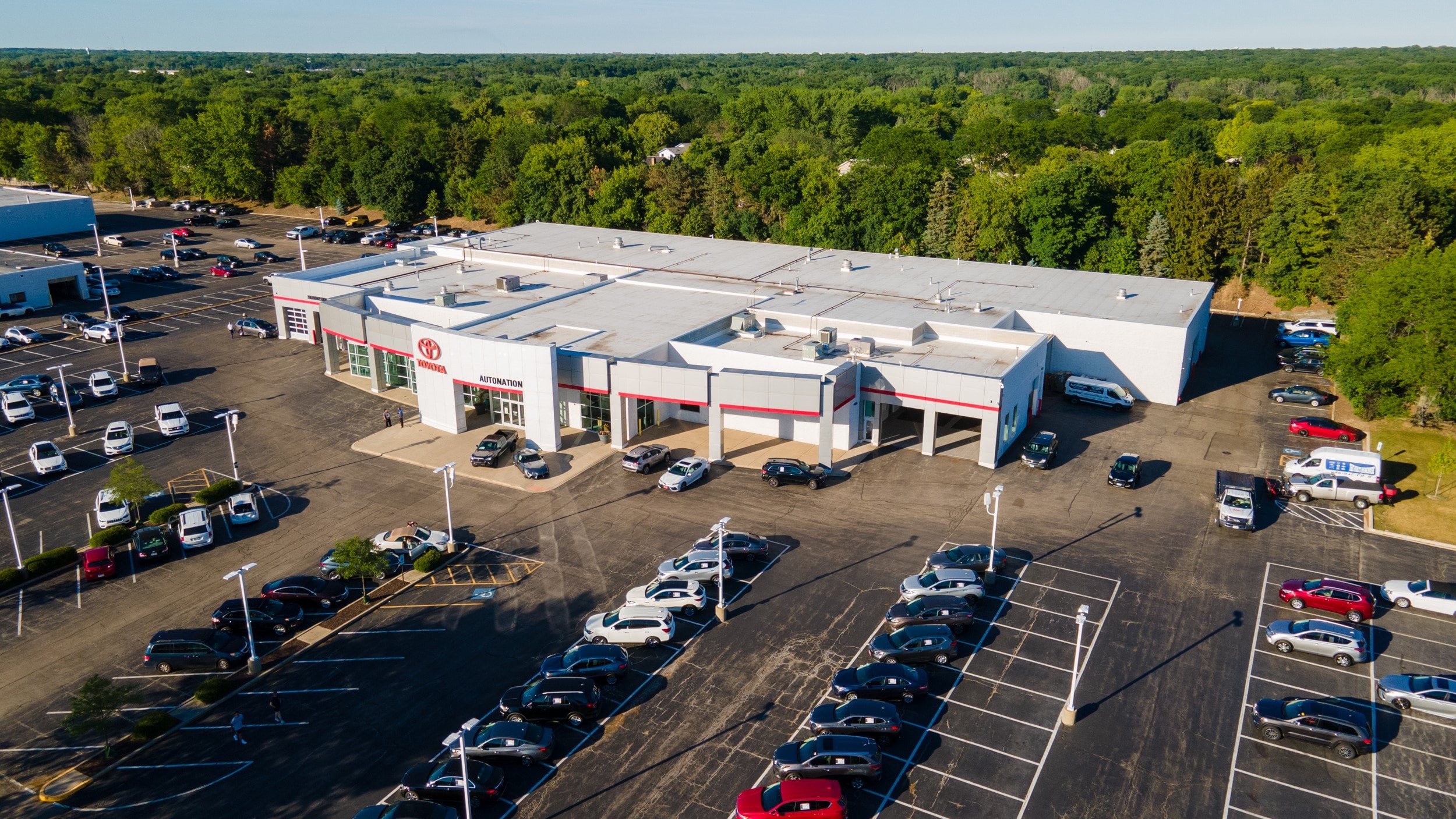 Overhead exterior view of AutoNation Toyota Libertyville. The building is grey and white and has some large windows. Many vehicles can be seen parked near the building, which is in a forested area.