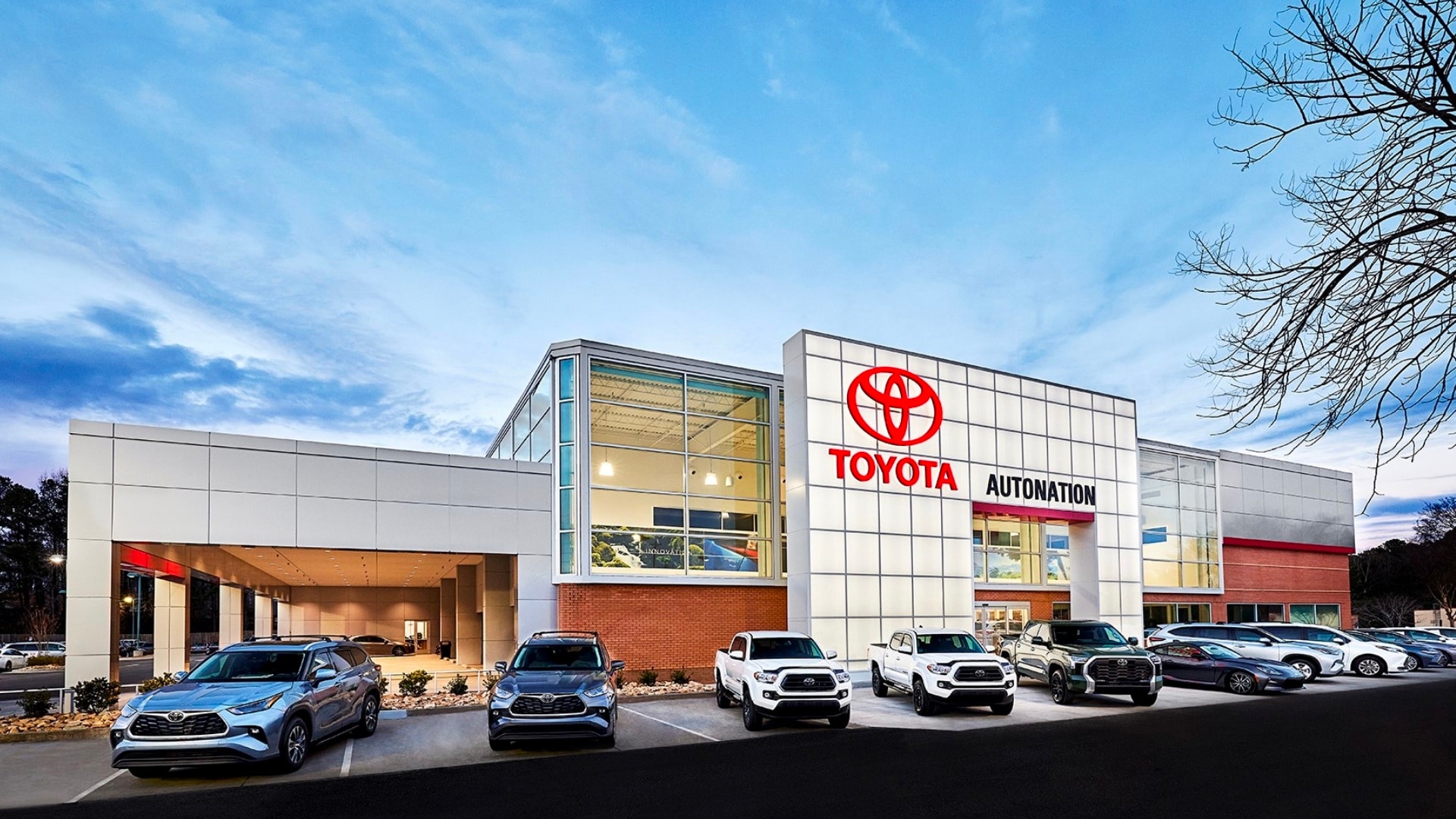 Exterior view of AutoNation Toyota Mall of Georgia. The building is grey and white and has some large windows. Many vehicles are seen parked lined up the building, which also has some trees nearby.