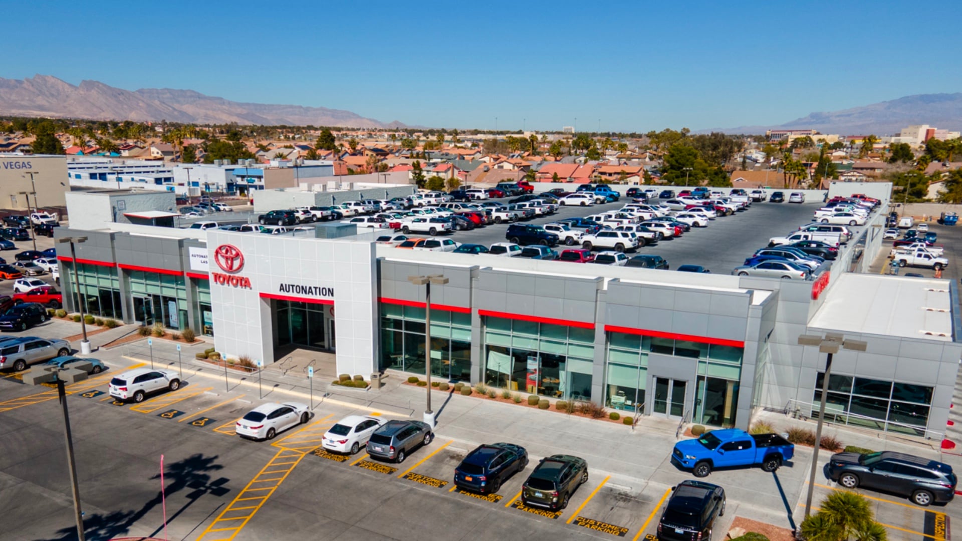 Overhead exterior view of AutoNation Toyota Las Vegas. The building is grey and white and has some large windows. Many vehicles can be seen parked near the building and on the roof. Mountains are visible in the distance.