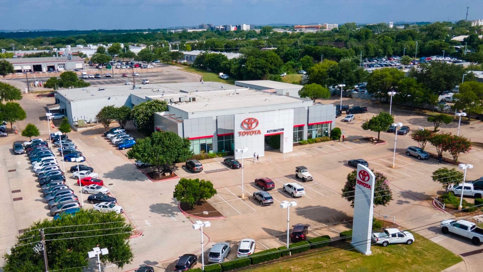 Overhead exterior view of AutoNation Toyota South Austin. The building is grey and white and has some large windows. Many vehicles are seen parked in rows near the building, which also has many trees nearby.