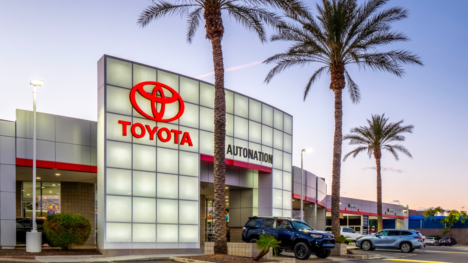 Exterior view of AutoNation Toyota Tempe. The building is grey and white and has some large windows. Several vehicles are seen near the building, which has several palm trees near it.