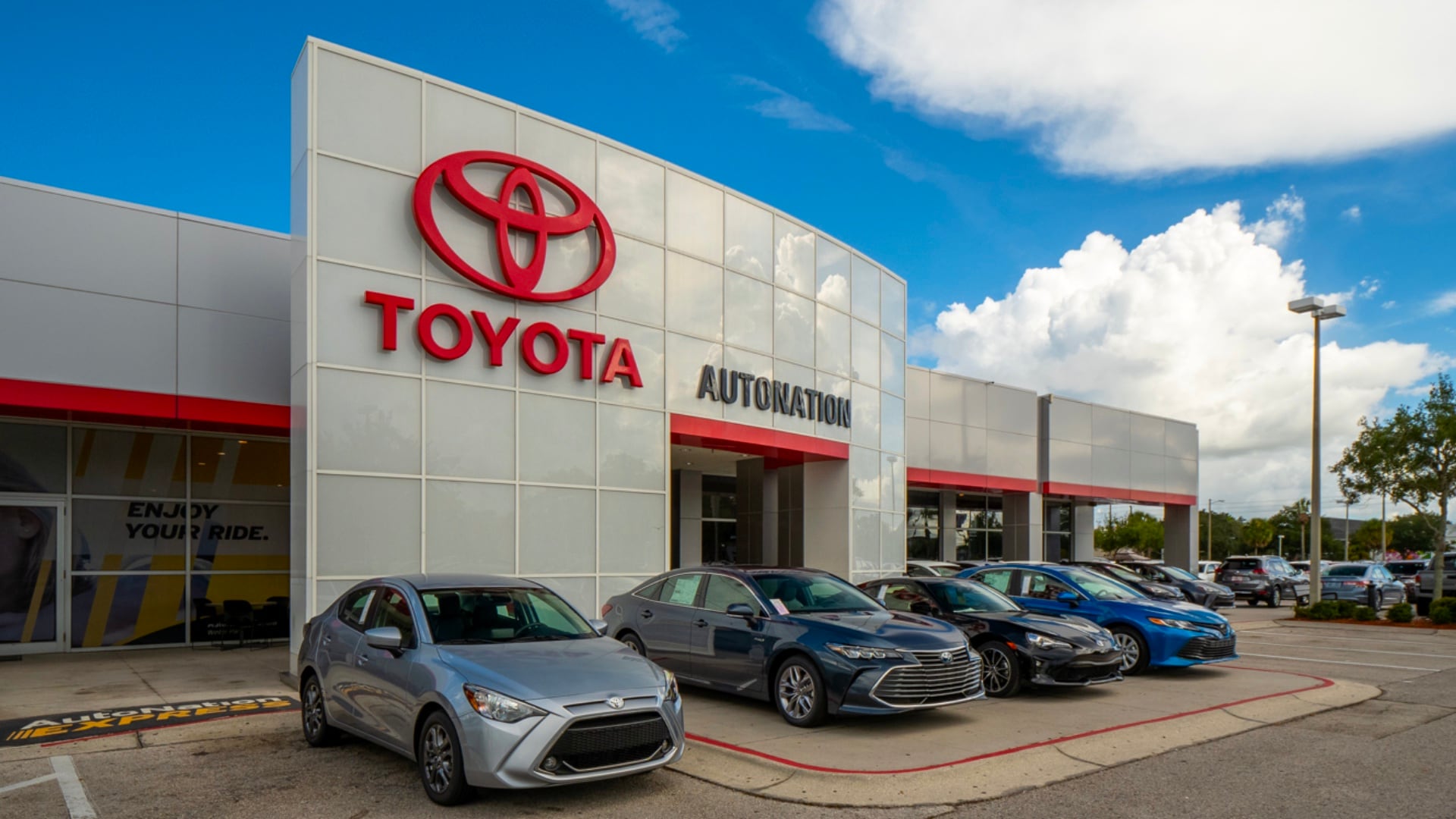 Exterior view of AutoNation Toyota Winter Park on a partly cloudy day. The building is grey and white and has some large windows. Several vehicles can be seen near the building, which also has some trees nearby.