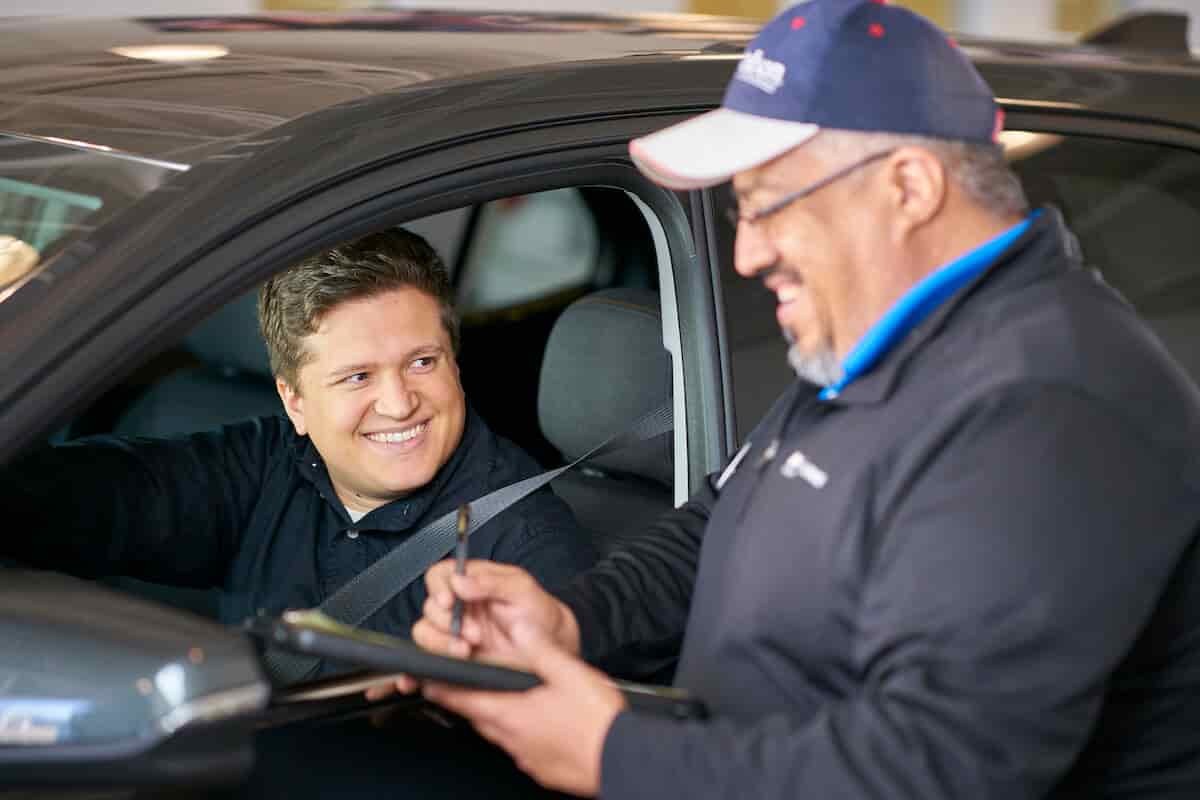 Man in a blue hat is laughing and holding a clipboard and pen while standing next to driver's side window of a vehicle. The vehicle's driver is smiling and looking at the man in the blue hat.