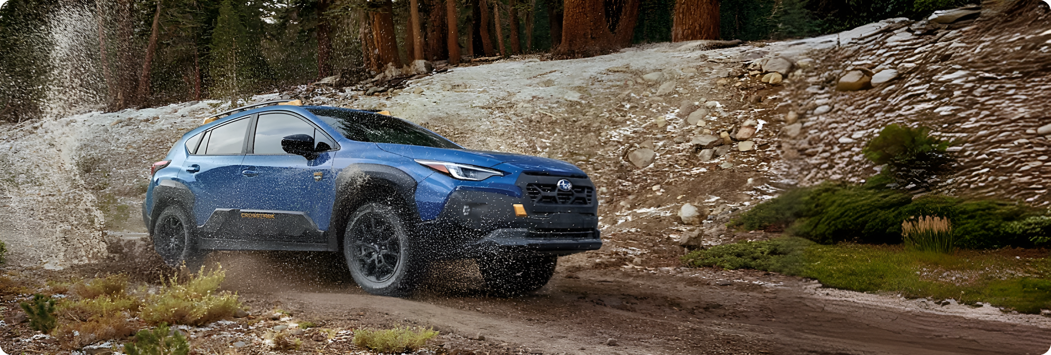 A blue Subaru Crosstrek throwing up water and mud as it climbs a rocky forest trail