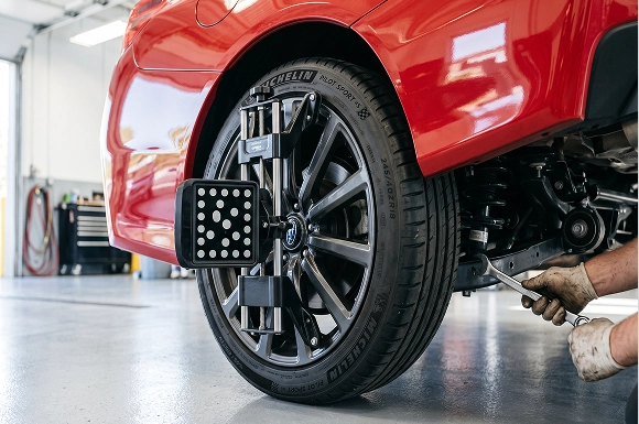 A close-up of an alignment target clamped to the front wheel of a red Subaru while a gloved Baldwin Subaru technician makes an adjustment underneath