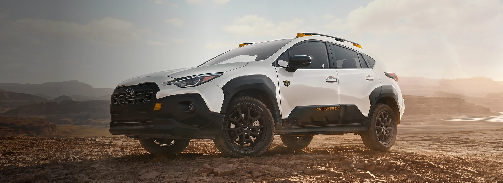 A white 2026 Subaru Crosstrek Wilderness with yellow roof-rail accents parked on red desert rock at sunset
