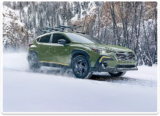 A green Subaru Crosstrek with a roof-mounted ski rack driving through deep snow past a birch forest