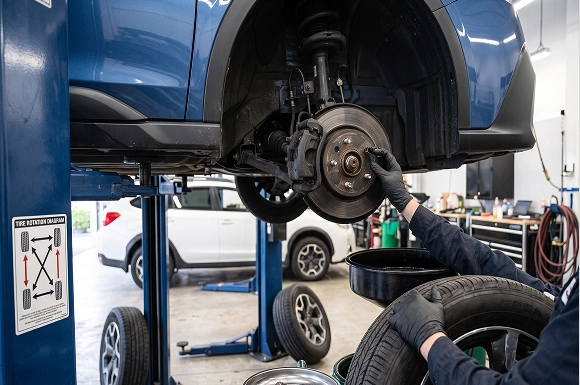A gloved Baldwin Subaru technician inspecting the exposed brake rotor and hub of a blue Subaru on a lift, with removed wheels stacked nearby and a tire rotation diagram posted on the lift column