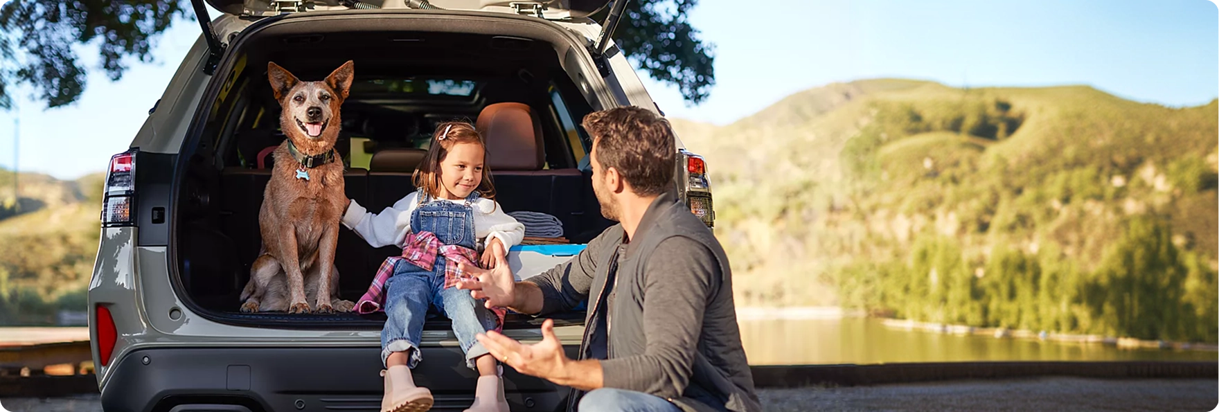 A dog and a young girl sitting in the open cargo area of a silver Subaru Forester while a man plays with them