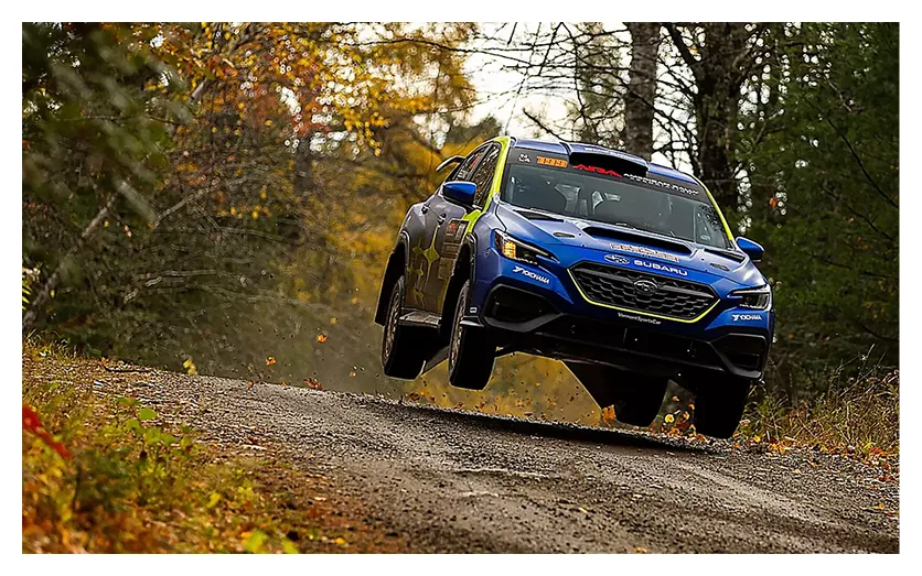 A blue Subaru WRX airborne over a rally stage crest