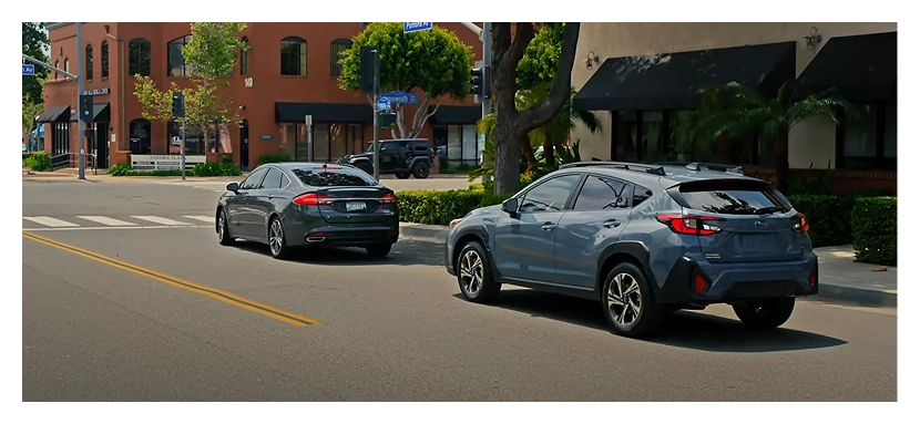 A teal Subaru Crosstrek slowing behind parked cars on a sunlit town street