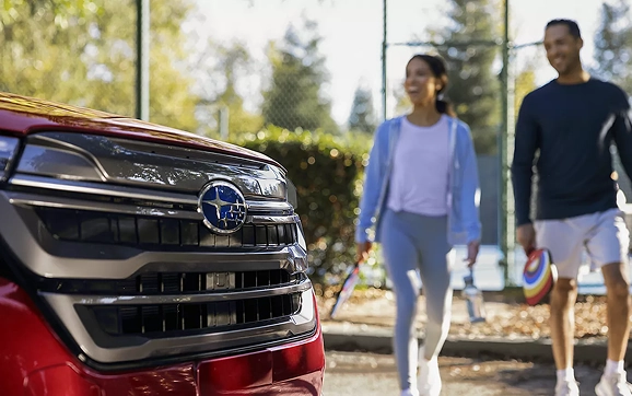 A couple walking past the front grille of a red Subaru Forester