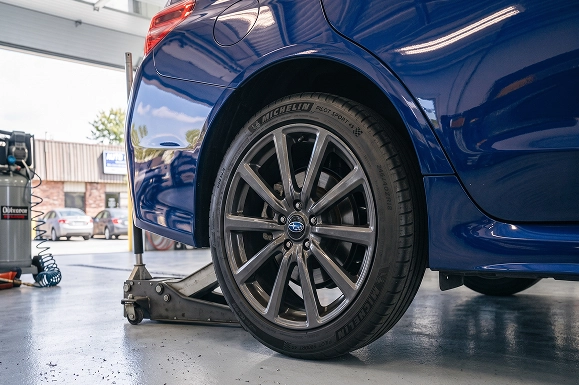 A close-up of the rear wheel and Michelin tire on a blue Subaru lifted on a jack in the Baldwin Subaru service bay