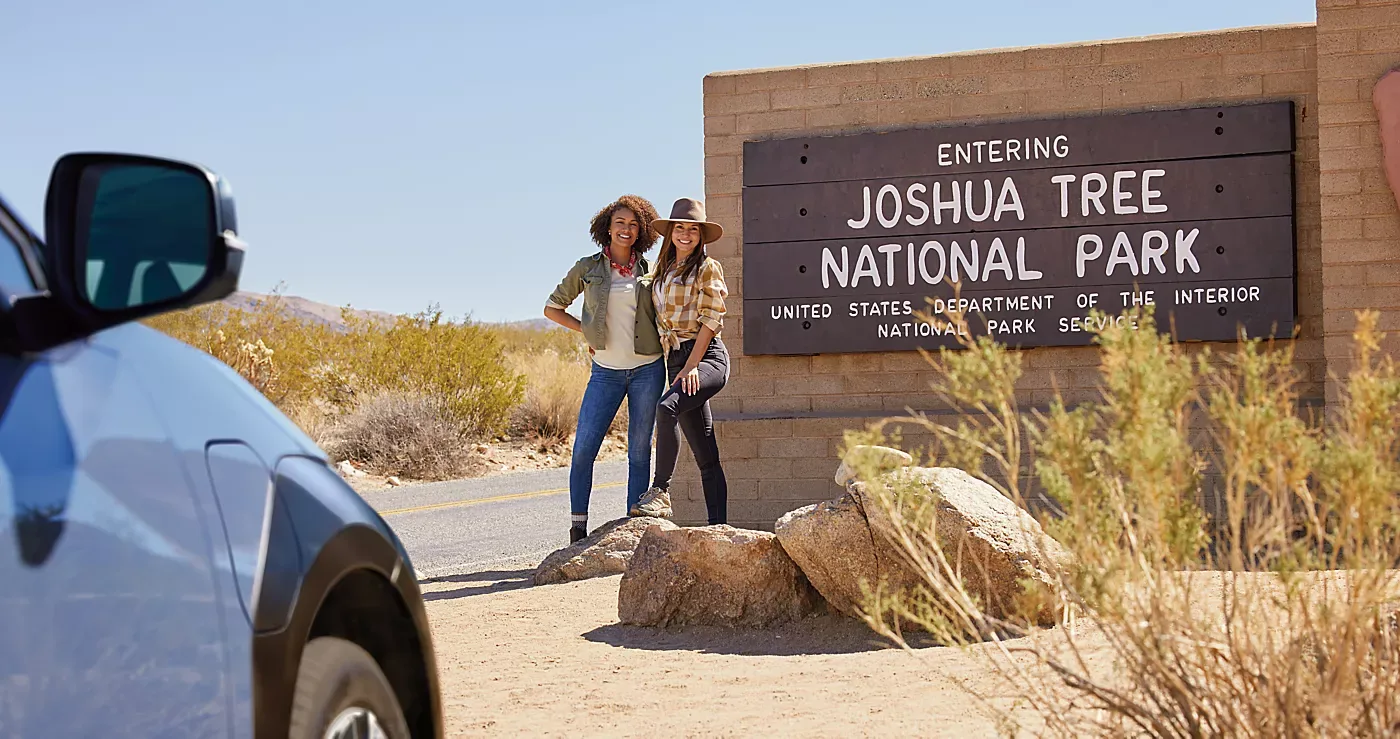 Hikers at the Joshua Tree National Park entrance sign beside a parked Subaru Solterra