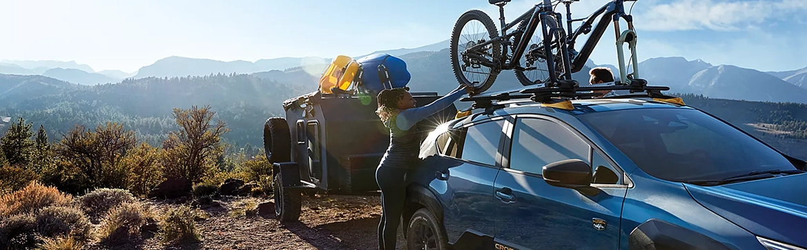 A person loading a mountain bike onto the roof rack of a blue Subaru Crosstrek at a mountain overlook