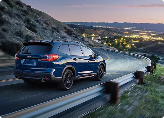 A blue Subaru Ascent on a mountain overlook at sunset with city lights below