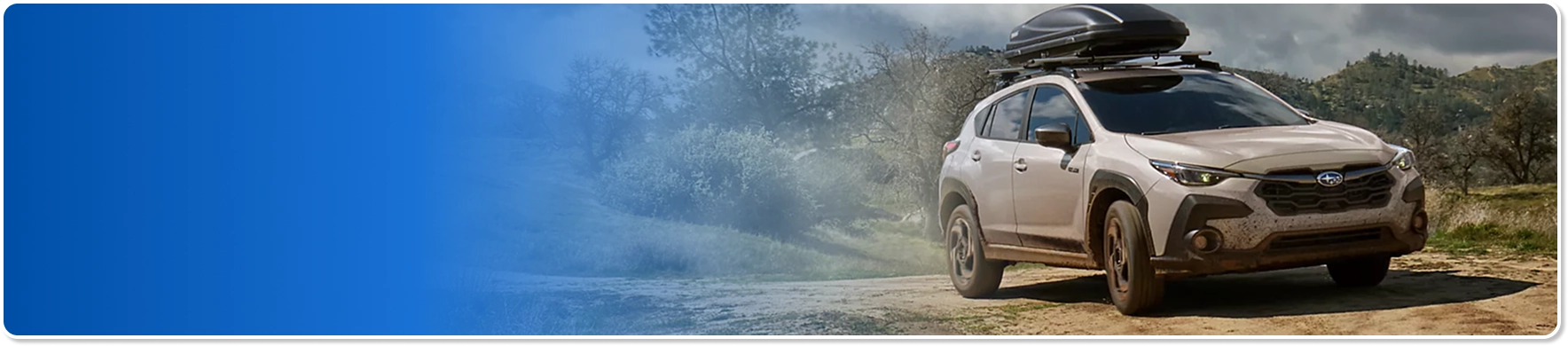 A champagne Subaru Crosstrek with a black roof cargo box driving a dusty trail past a misty hillside
