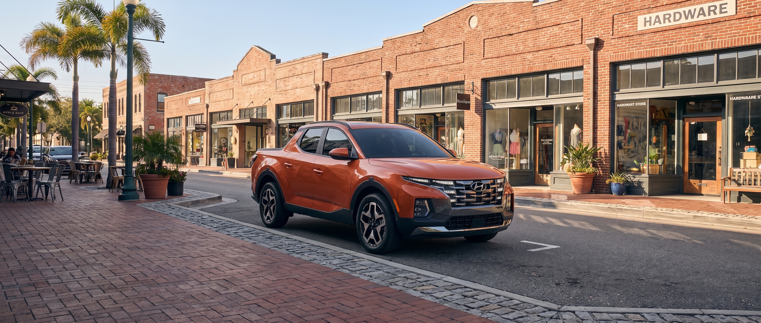 A 2026 Hyundai Santa Cruz in a warm Gulf Coast light, parked on a revitalized urban street with brick-accented sidewalks and a mix of small businesses visible in the background, evoking a downtown commercial district feel in summer.