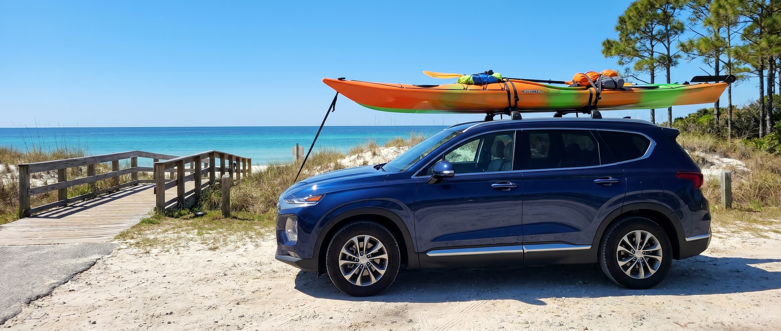 A loaded kayak strapped to the roof of a dark blue SUV parked near a sandy boat launch at St. Andrews State Park, with calm turquoise water and a clear Florida sky in the background.