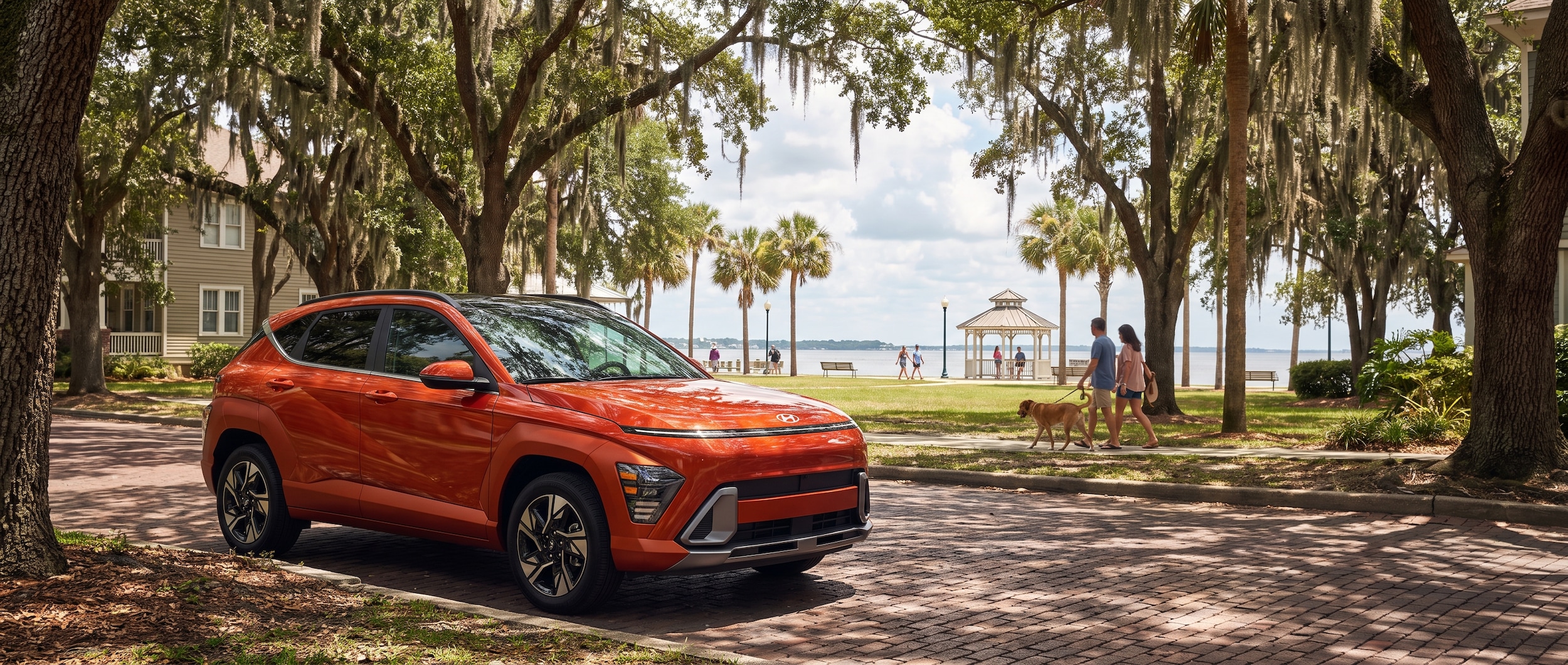 A 2026 Hyundai Kona in a bold exterior color parked on a shaded residential street in a Gulf Coast neighborhood, with a waterfront park visible in the warm summer background, suggesting everyday Panama City, FL life