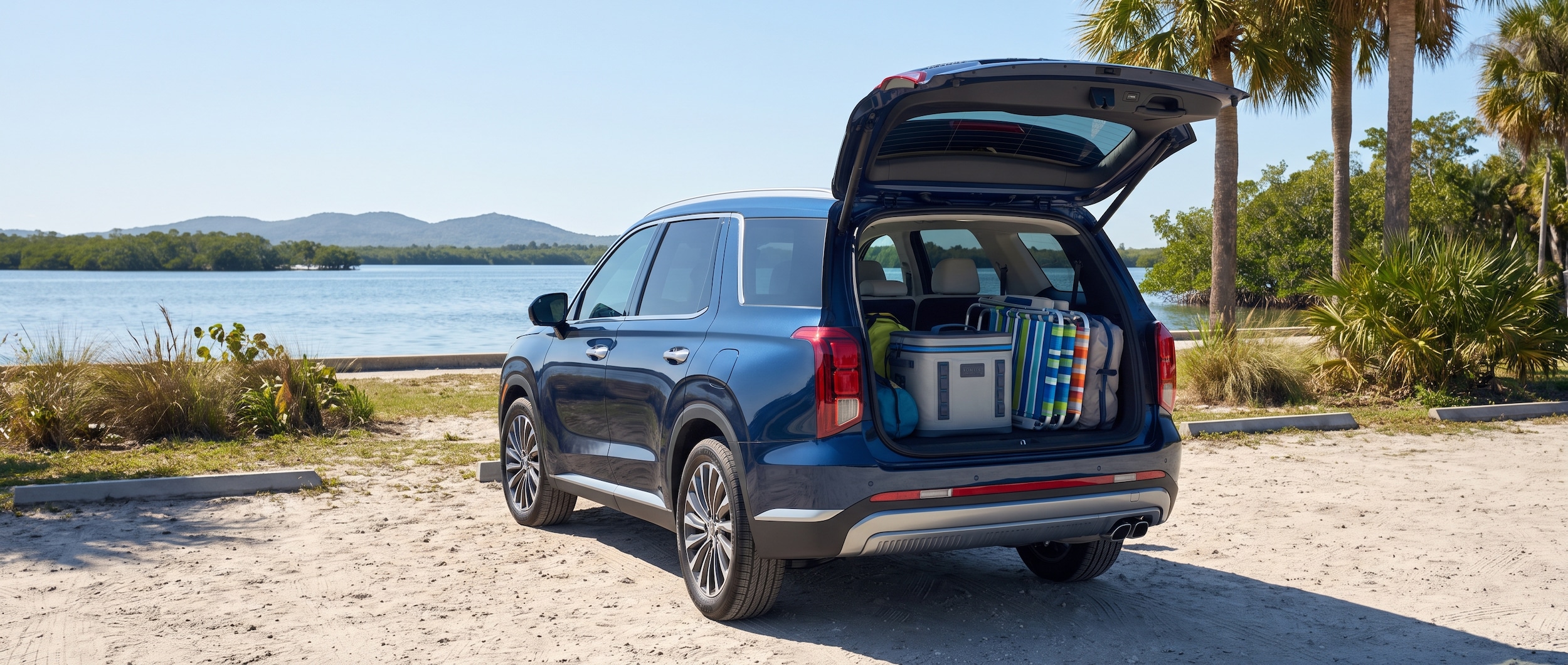 A 2026 Hyundai Palisade Hybrid in a deep blue or forest green, parked at a waterfront park with a North Bay backdrop on a bright Florida summer day, rear liftgate open revealing loaded gear including a soft cooler and beach chairs
