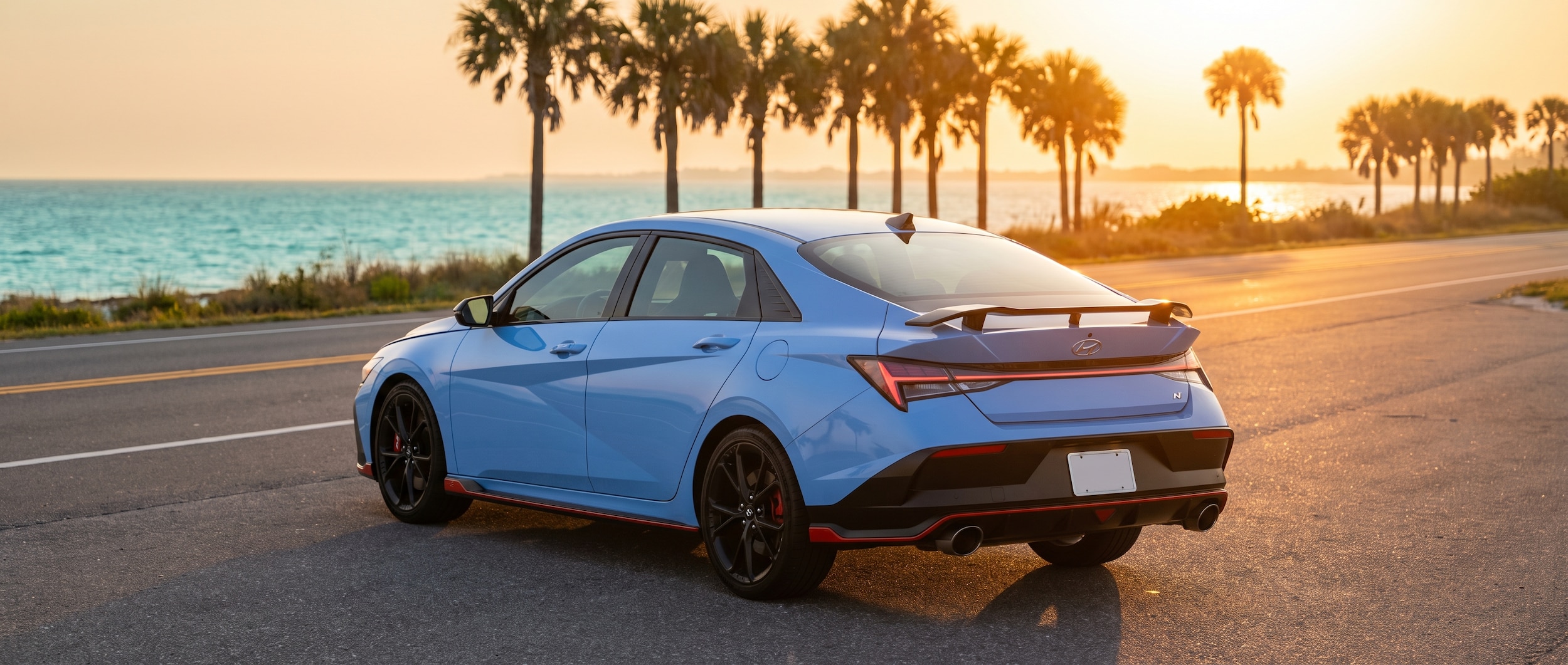 A 2026 Hyundai Elantra N in Performance Blue parked along a Gulf-side strip road at golden hour, palm trees and turquoise water visible in the background, rear wing and dual exhaust outlets prominent, summer Florida coastal setting