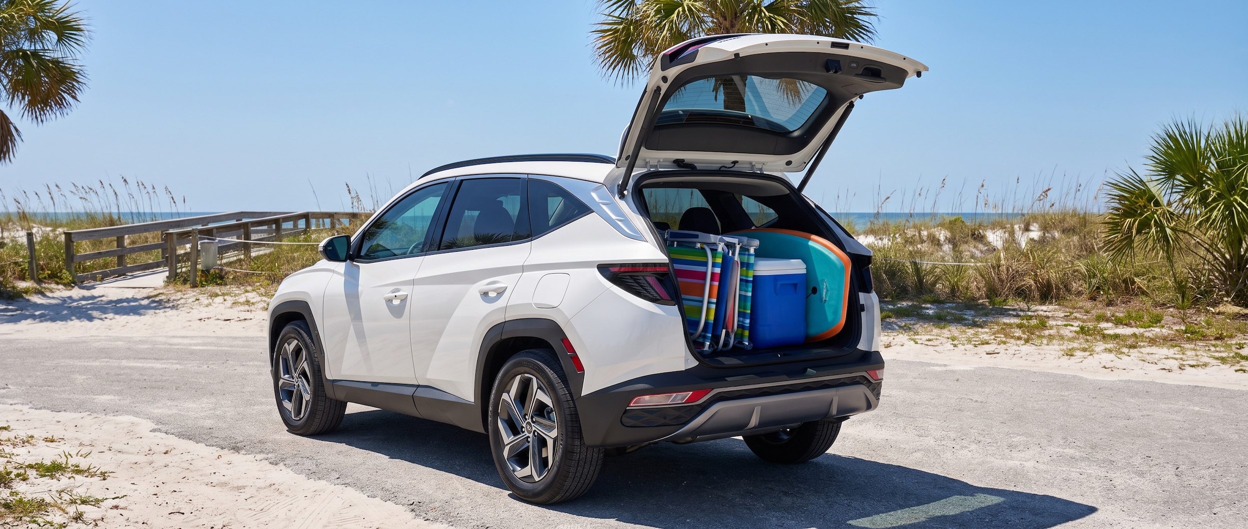 A 2026 Hyundai Tucson Hybrid in Abyss Black or Serenity White parked near a Florida Gulf Coast beach access road in bright July sunlight, with an open rear cargo area loaded with beach chairs, a cooler, and boogie boards, blue sky and palm fronds visible in the background