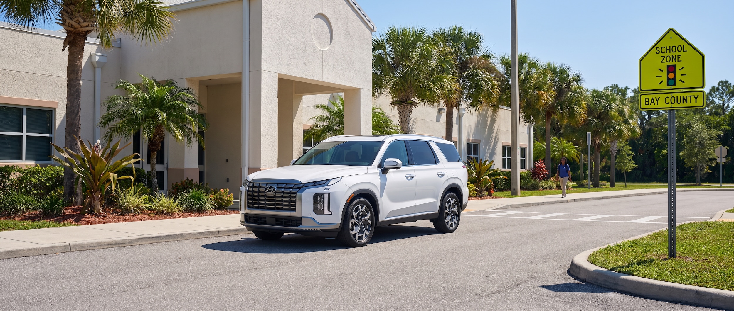 A 2026 Hyundai Palisade in white parked outside a Florida school entrance on a bright summer morning, with a Bay County school zone sign visible in the background. The vehicle's rear door is open, showing the spacious interior and three rows of seating.