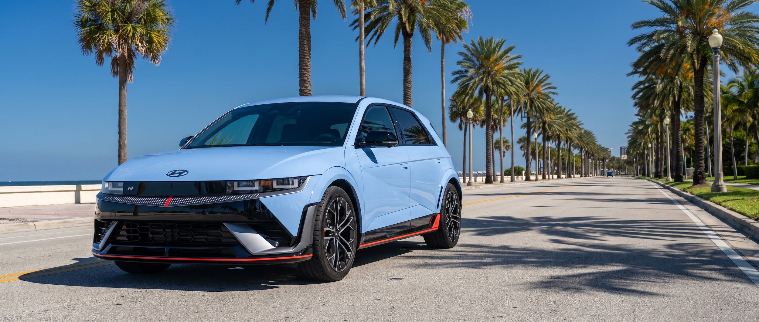 2026 Hyundai IONIQ 5 N in Performance Blue Pearl parked along a wide, sun-drenched multi-lane boulevard with Gulf Coast palm trees and deep blue summer sky, front three-quarter angle showing the aggressive mesh grille, red accent stripe, and 21-inch forged wheels