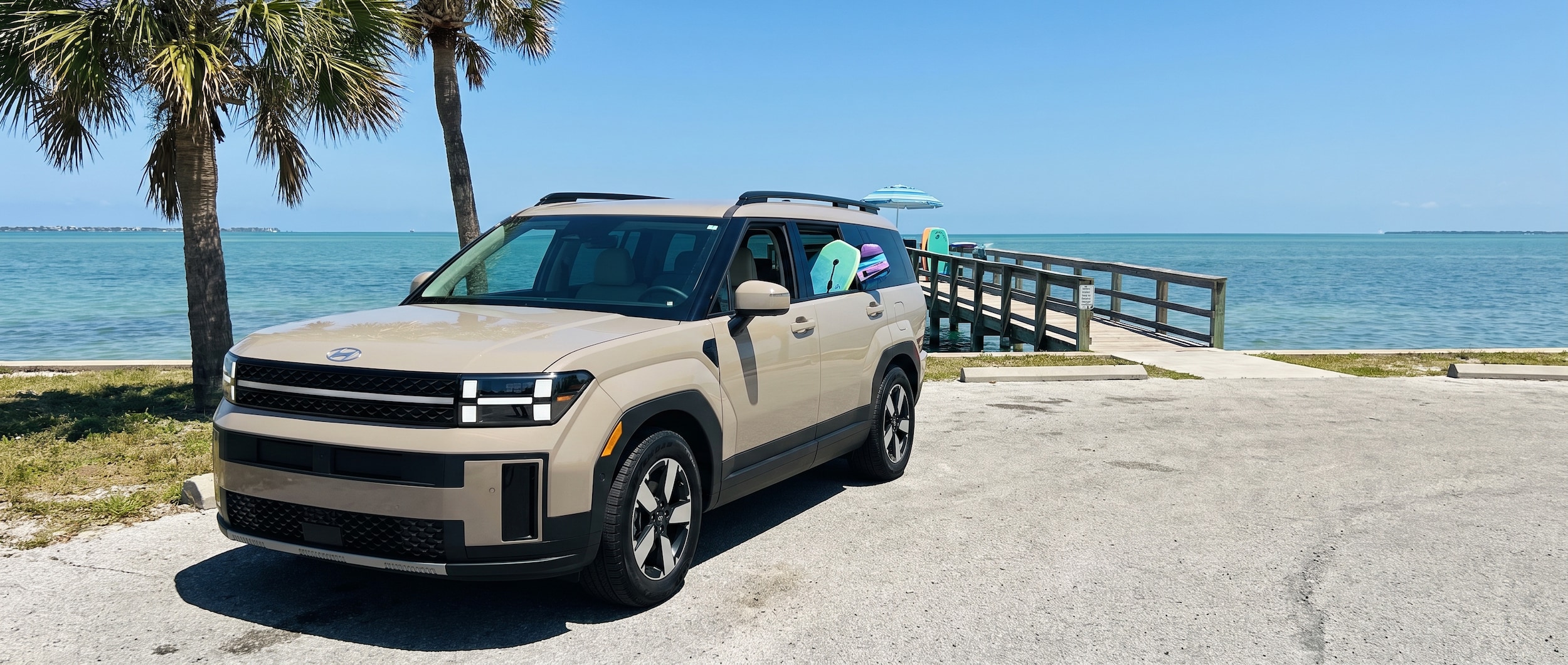 A 2026 Hyundai Santa Fe in a coastal Florida setting, parked near a waterfront with bright Gulf Coast summer light, lush palm trees, and a clear blue sky. The SUV faces forward showing its bold squared-off front end and H-shaped LED lighting. Family gear is visible through the rear window, suggesting a loaded school run or beach day outing.