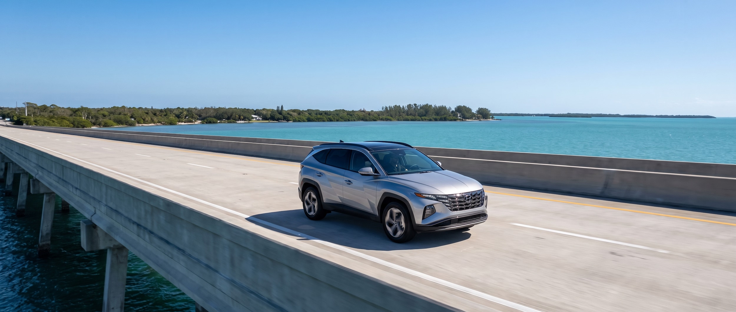 A 2026 Hyundai Tucson Hybrid in Shimmering Silver driving across a wide concrete highway bridge over a blue coastal bay, bright Florida summer sky, lush green mainland visible in the background
