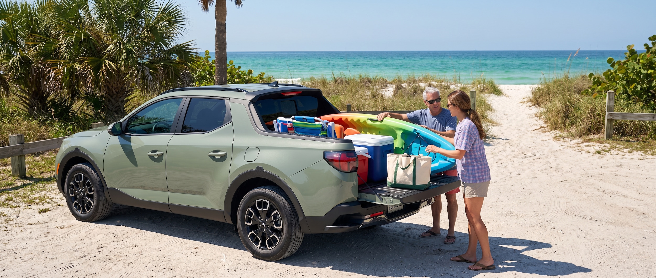 A 2026 Hyundai Santa Cruz in a coastal Florida setting, tailgate down, kayaks and beach gear being loaded in the bed on a bright summer morning, emerald green water visible in the background
